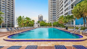 Una piscina rodeada de sillas de descanso y palmeras, con edificios altos al fondo.