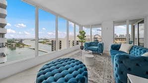 Una sala de estar moderna con ventanas amplias, un sofá azul, un otoman redondo y una planta en maceta.