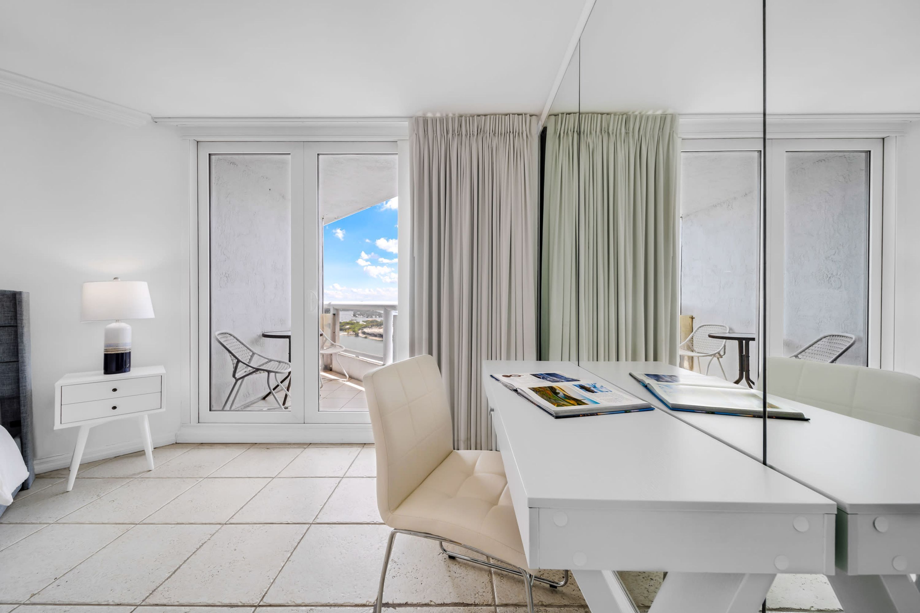 Habitación de hotel moderna con un escritorio blanco, una silla, una mesita de noche con lámpara y vistas al balcón.