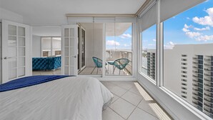 Un dormitorio con una cama grande, una pared de acento azul y un balcón con sillas y vistas a la ciudad.