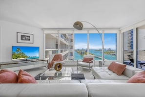 A modern living room with a large TV, comfortable seating, and a view of a cityscape and water.