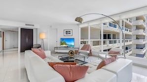 A modern living room with a white sofa, a coffee table, and a TV. There is a balcony with white railings and a view of a building. - Highrise Charm (Miami)