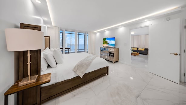 Quarto moderno com uma cama grande, uma mesa de cabeceira, uma televisão e vista para o exterior por grandes janelas.