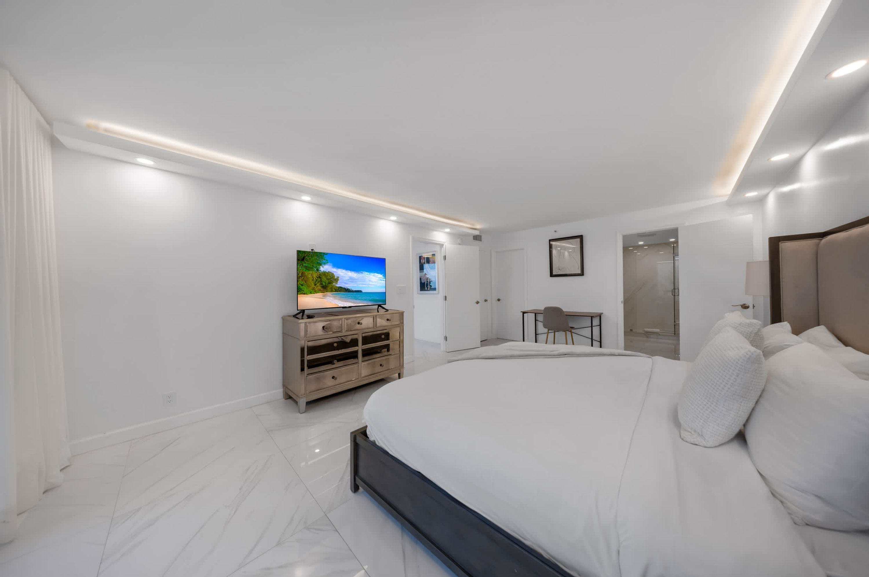 Une chambre à coucher moderne avec un grand lit, un téléviseur posé sur une commode, un bureau et un dressing.