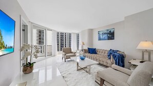 A modern living room with a sofa, coffee table, and a TV. There is a large window with a view of a cityscape. - Azure Skies High Floor (Miami)