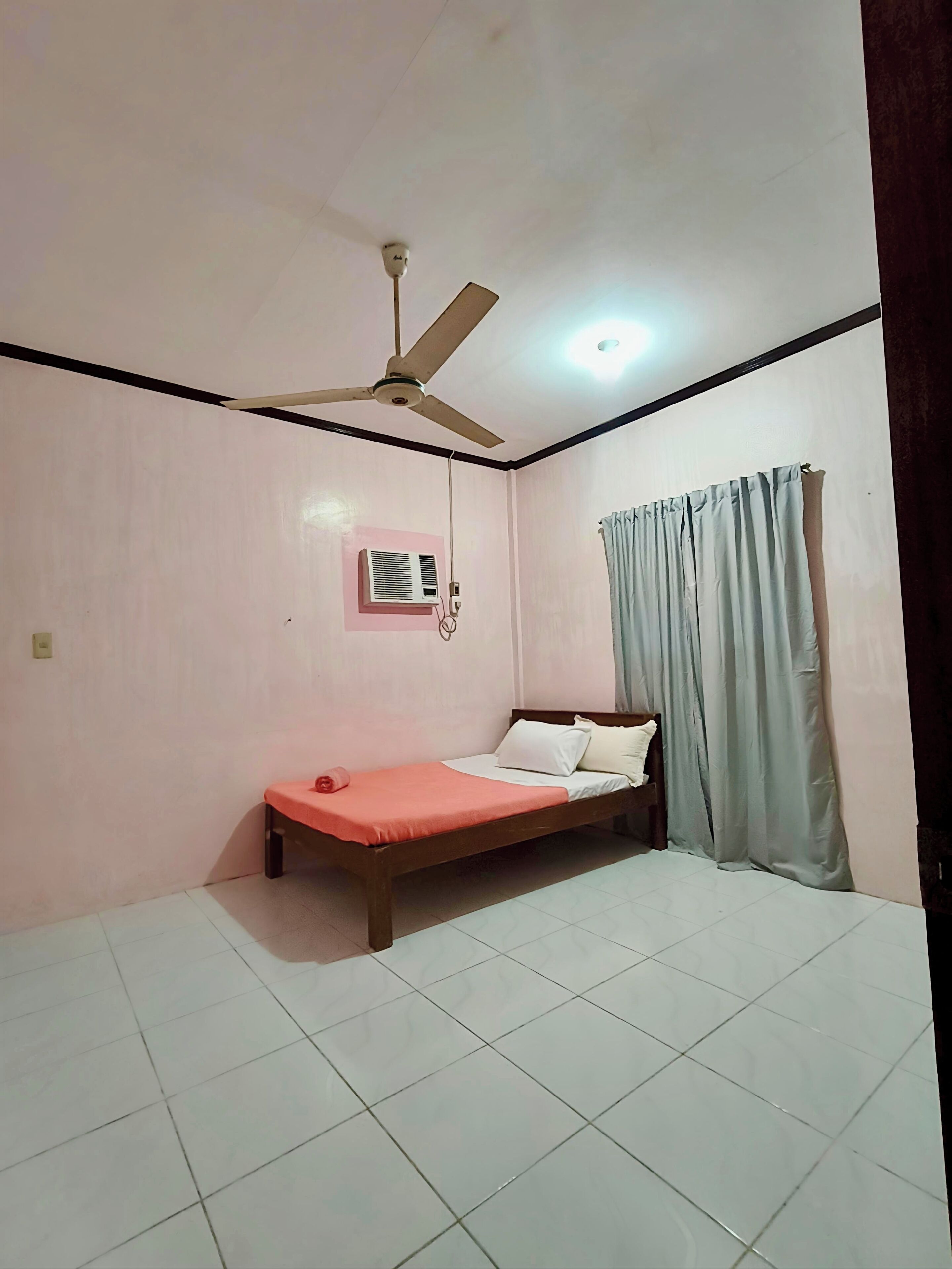A room with a bed, a ceiling fan, an air conditioning unit, and a window with curtains.