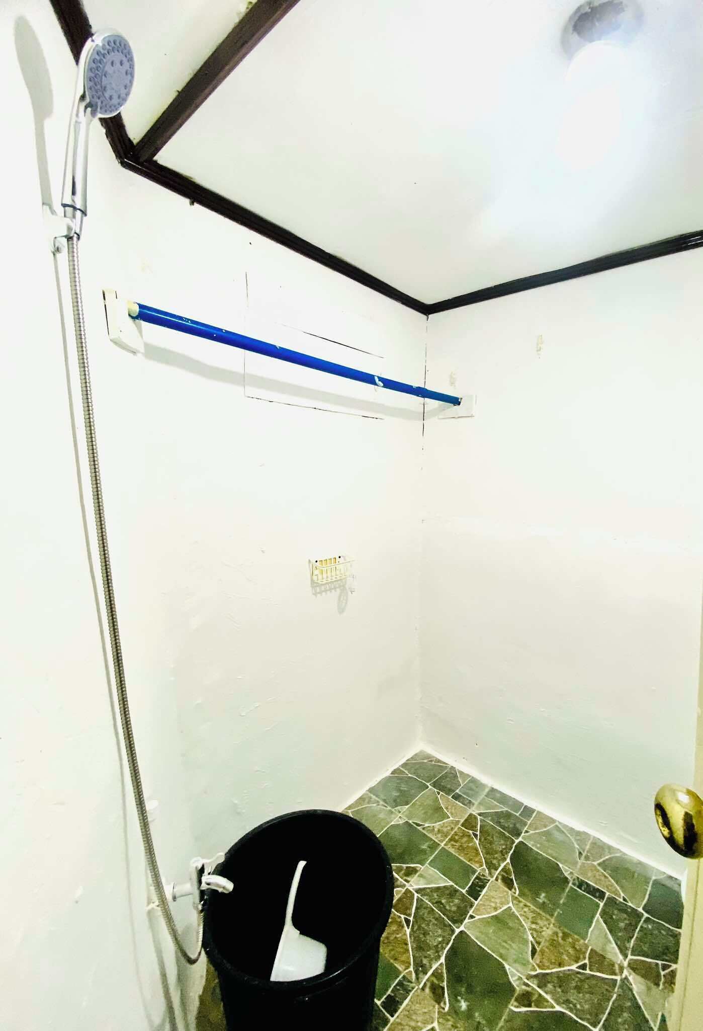 A small, enclosed shower area with a showerhead, a blue towel rack, and a black bucket on the floor.