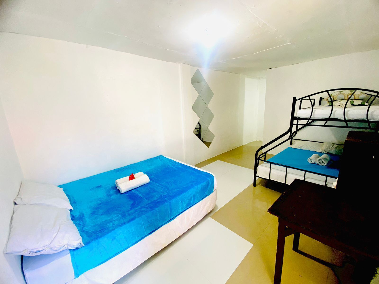 Une petite chambre avec un lit simple, un matelas bleu, des oreillers blancs et une rose rouge. On aperçoit un deuxième lit, également avec un matelas bleu et des oreillers blancs, au fond de la pièce.