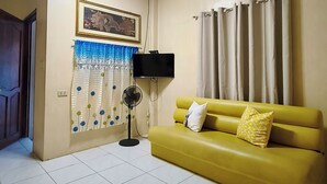 Un soggiorno con un divano giallo, una ventola nera, un televisore e un quadro appeso al muro.