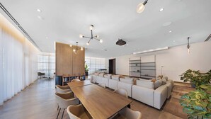 A modern dining area with a long wooden table and beige chairs, a sofa set, and a large window with sheer curtains.