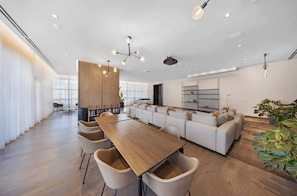 Un comedor moderno con una larga mesa de madera y sillas color beige, un conjunto de sofá y un amplio ventanal con cortinas translúcidas.