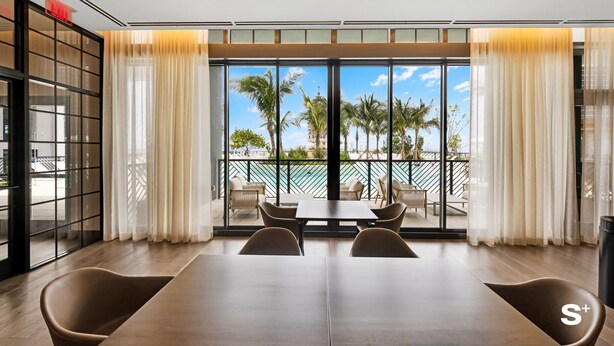 A modern dining area with a large table and chairs, large windows with curtains, and a view of palm trees and a pool.
