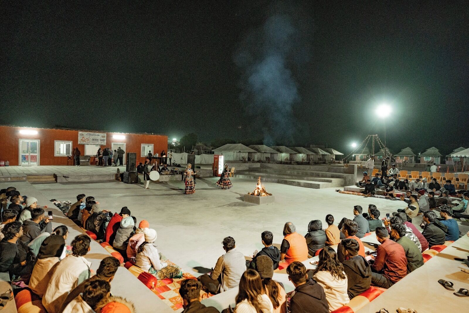 An outdoor gathering with a central fire, people seated on mats, and tents in the background.