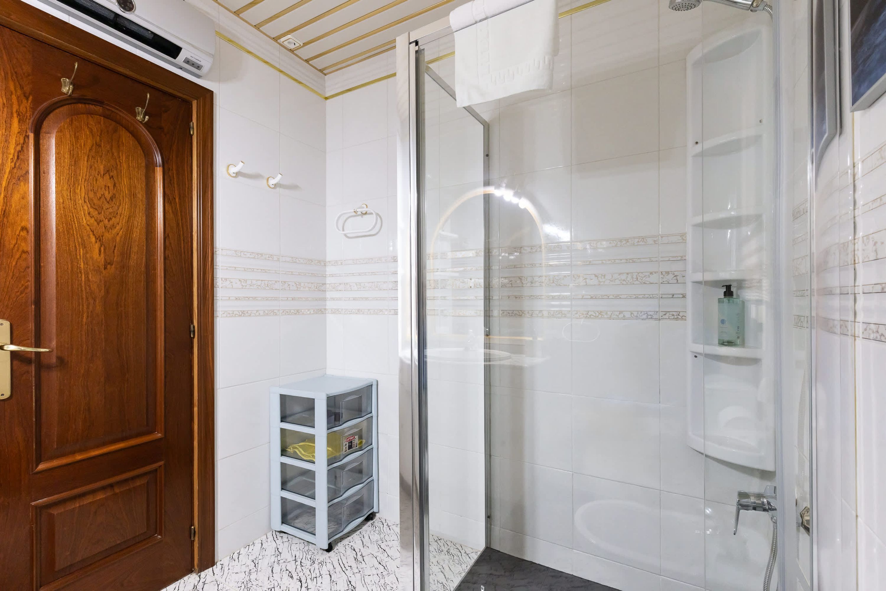 Une salle de bain avec une porte en bois, un espace de douche en verre et un mur recouvert de carreaux blancs agrémenté d’une bordure décorative.