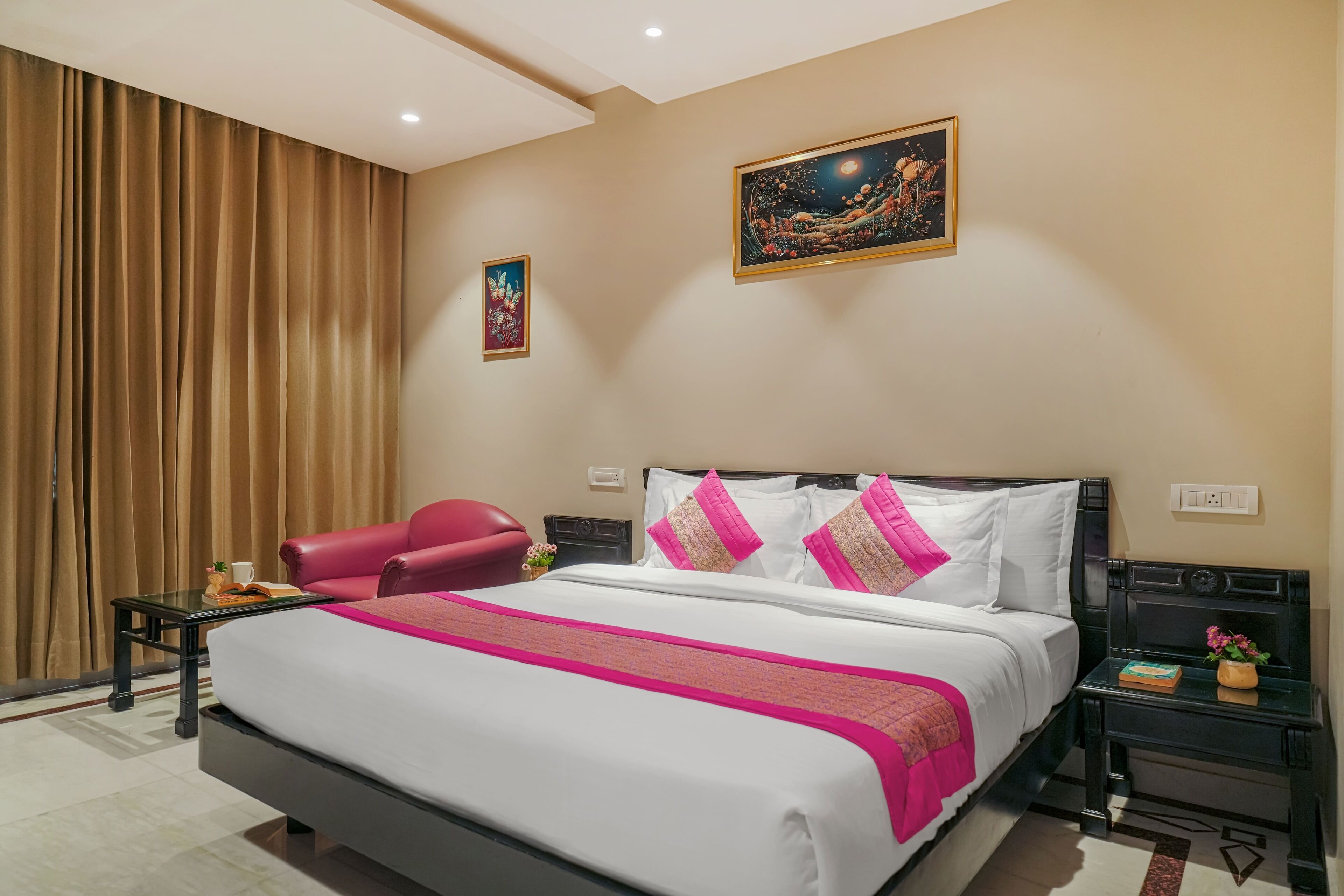 A hotel room with a large bed, a red chair, a small table with a book and a cup, and a nightstand with a plant.