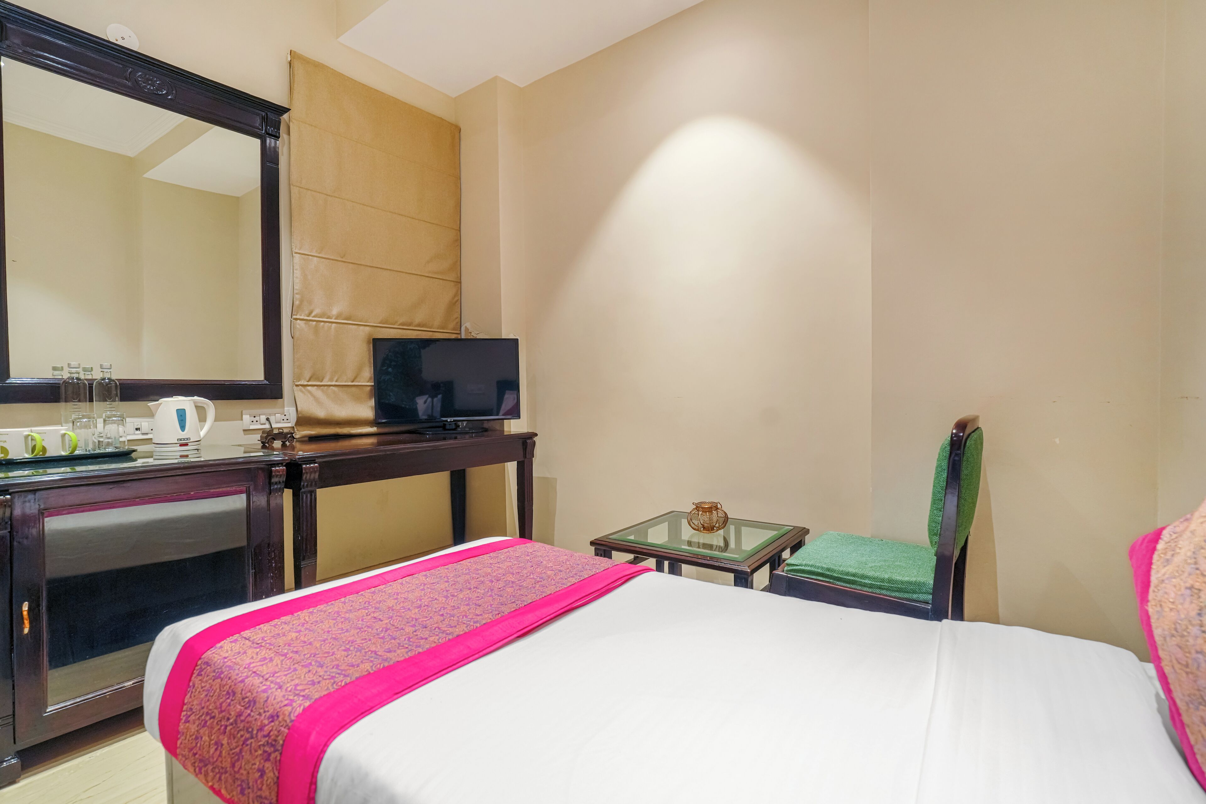 Habitación de hotel con cama, escritorio con televisor, espejo, mesita de noche con lámpara y una silla.