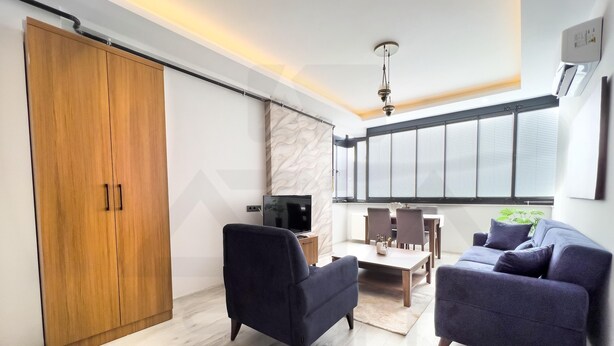 A modern living room with a sofa, a coffee table, and a TV. There's a wooden wardrobe and a window with blinds. - ARİA SUİT KONAKLAMA (Mezitli)