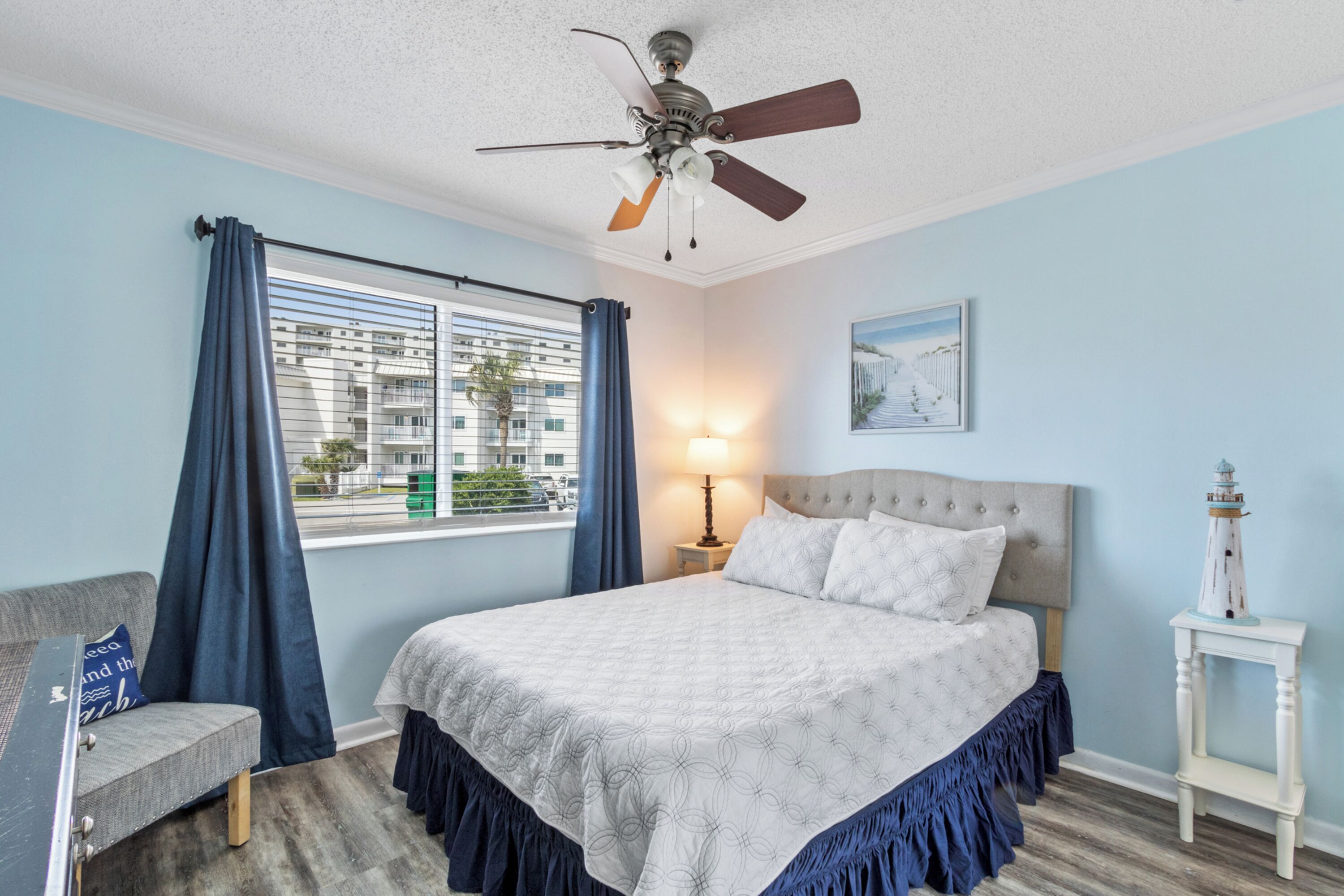 A bedroom with a bed, a ceiling fan, a nightstand with a lamp, a chair, and a window with curtains.