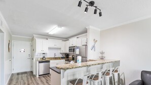 Een moderne keuken met witte kasten, een granieten werkblad en roestvrijstalen apparatuur. Drie barkrukken en een zwarte lederen stoel.