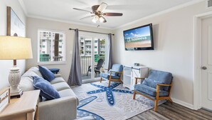 Een woonkamer met een bank, stoelen, een zeesterren-tapijt, een plafondventilator, een televisie en uitzicht op een balkon met gebouwen.