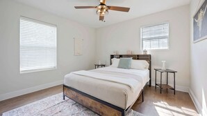 Ein Schlafzimmer mit einem Bett, einem Deckenventilator, zwei Nachttischen und einem Fenster mit Jalousien.