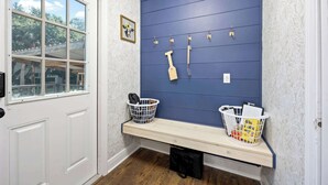 Ein Zimmer mit weißer Tür, einem Fenster mit Aussicht, einer blauen Wand mit Haken, einer Bank mit Körben und einer Wand mit Muster.