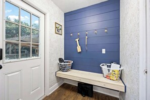 A room with a white door, a window with a view, a blue wall with hooks, a bench with baskets, and a patterned wall.