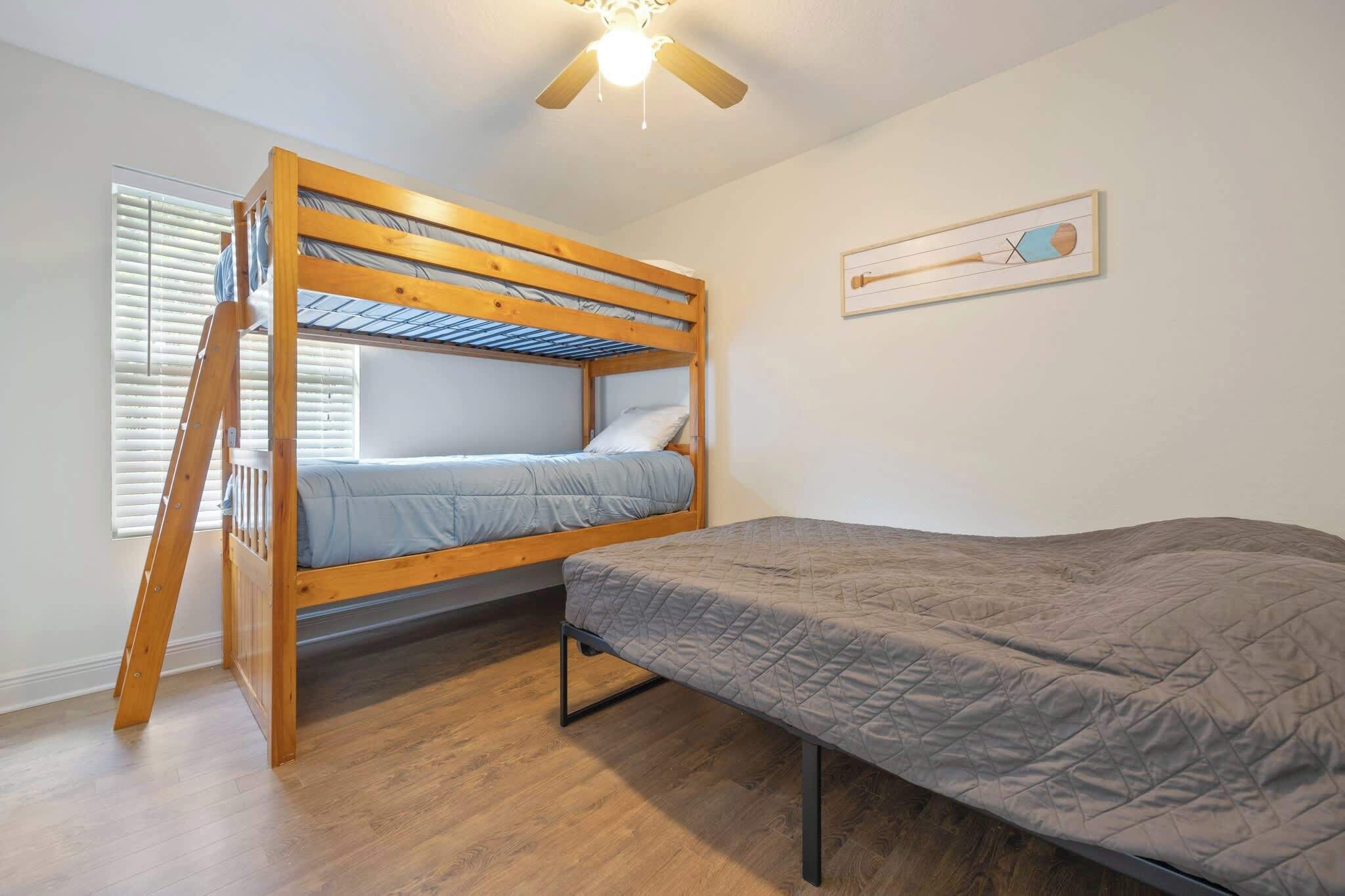 Ein Stockbett mit einer Leiter, ein Einzelbett, ein Deckenventilator, ein Fenster mit Jalousien und ein gerahmtes Bild an der Wand.