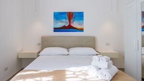 Une chambre d’hôtel moderne avec un grand lit, du linge de lit blanc, une tête de lit, des tables de chevet et un tableau représentant un volcan.