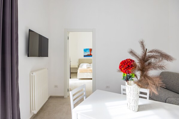 A room with a white table, a white chair, a white radiator, a grey sofa, and a TV mounted on the wall. - Maison Vittorio Emanuele (Naples)