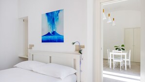 A bedroom with a bed, a white headboard, a wall-mounted shower, a dining area with a table and chairs, and a painting of a blue landscape. - Maison Vittorio Emanuele (Naples)