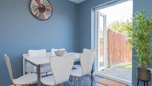 A dining area with a white table and chairs, a clock on the wall, and a potted plant. - Stylish 3 bedroom house with parking and private garden. Ideal for families, groups or work stays. (Cheltenham)