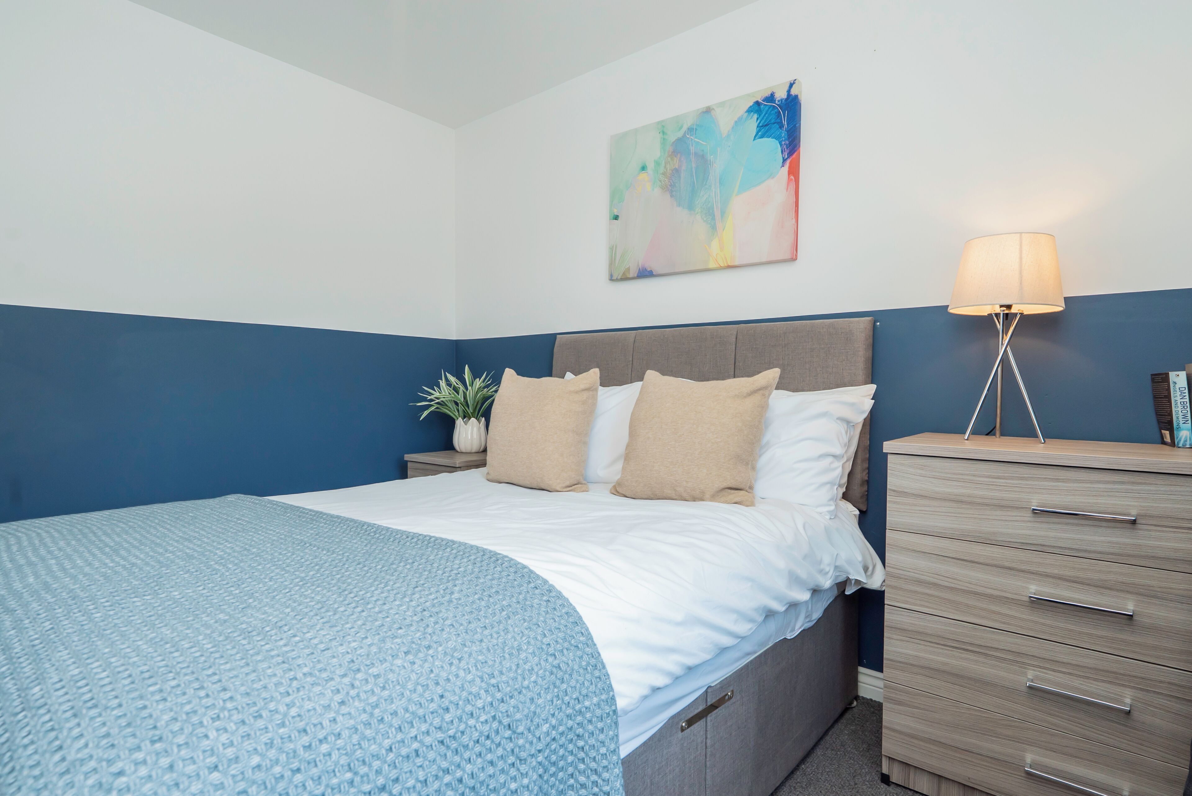 A bedroom with a bed, bedside table, lamp, and a wall-mounted abstract painting.