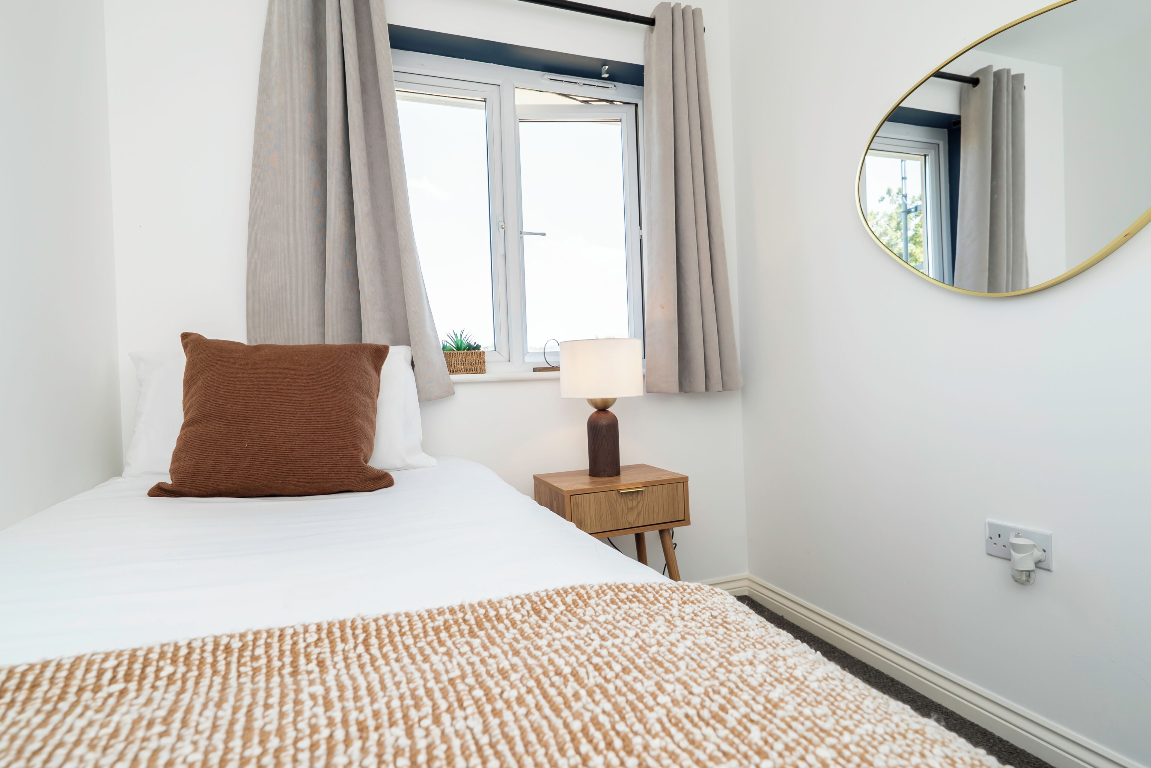 Un dormitorio con cama, mesita de noche con lámpara, ventana con cortinas y espejo colgado en la pared.