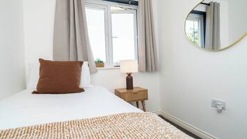 Un dormitorio con cama, mesita de noche con lámpara, ventana con cortinas y espejo colgado en la pared.