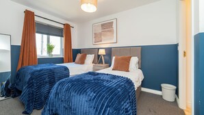 A hotel room with two beds, a window with curtains, a painting on the wall, and a lamp on the nightstand. - Stylish 3 bedroom house with parking and private garden. Ideal for families, groups or work stays. (Cheltenham)