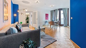 A modern living room with a grey sofa, a dining area with a table and chairs, and a kitchen visible through an open door. - 4ty3-Apartments Naschmarkt City Apt (Vienna)