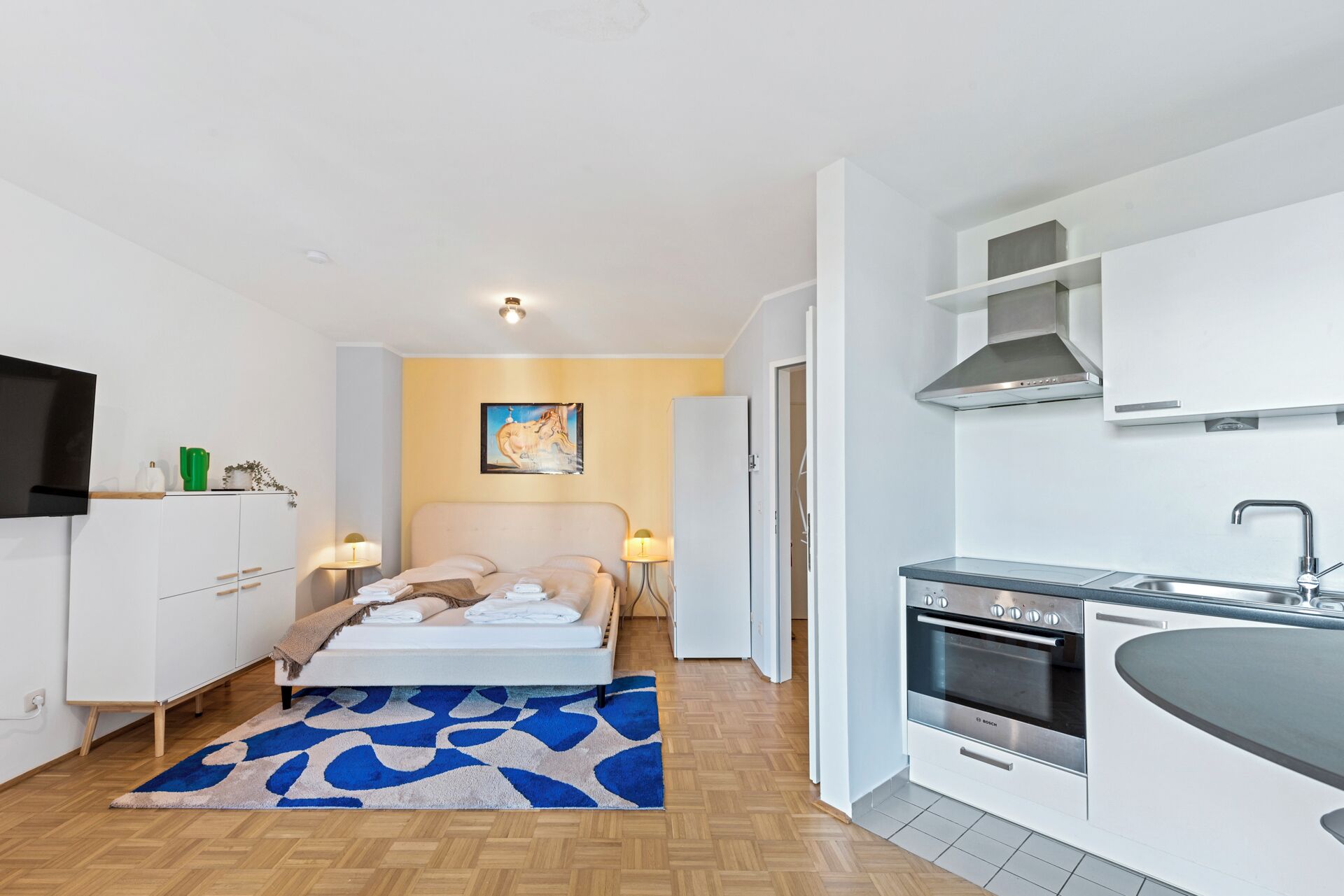 Une chambre d’hôtel compacte comprenant un lit, des lampes de chevet, une petite table, un canapé, un coin cuisine avec une cuisinière et un évier, et un tableau au mur.