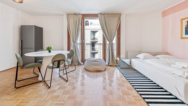 A hotel room with a bed, a dining table with chairs, a small kitchen area, and a view of a building through the window. - 4ty3-Apartments Naschmarkt City Apt (Vienna)