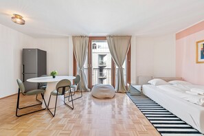 Ein Hotelzimmer mit Bett, Esstisch und Stühlen, einer kleinen Küchenzeile und Blick auf ein Gebäude durch das Fenster.