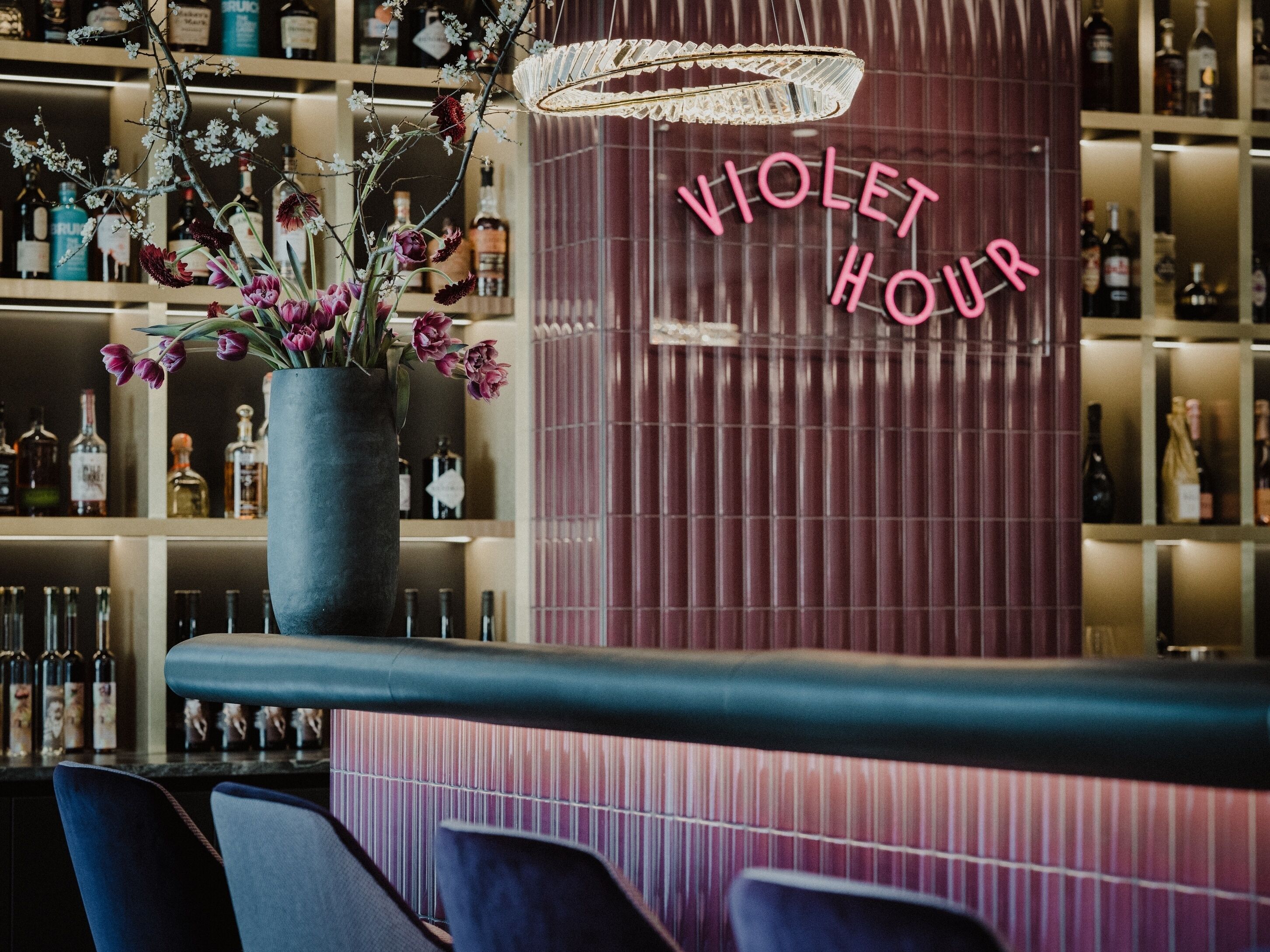 Et barområde med et neonskilt der siger "VIOLET HOUR" og en stor potteplante med blomster.