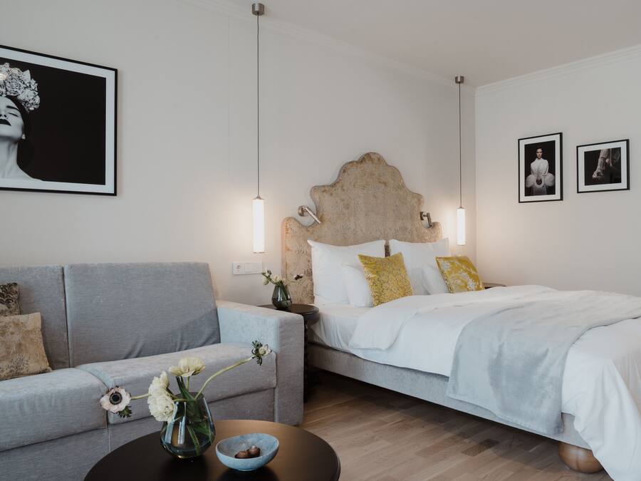 Un dormitorio moderno con un sofá gris, una cama con ropa de cama blanca, un florero con flores y dos luces fijadas a la pared.