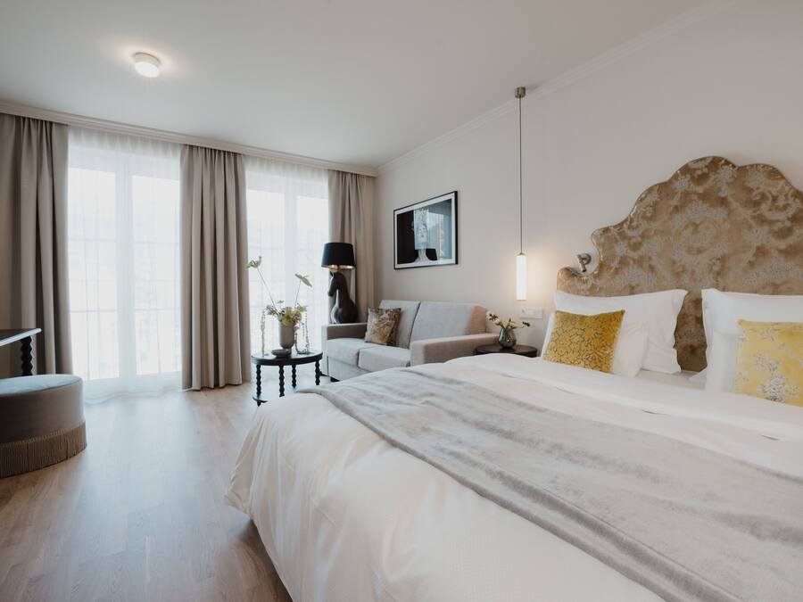Una camera d'hotel moderna con un letto grande, un divano, un tavolino con una lampada e un'ampia finestra con tende.