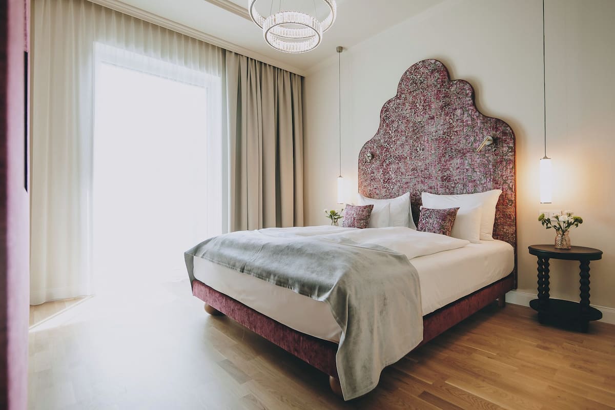 A neatly made bed with a floral headboard, a bedside table with a lamp, and a large window with curtains.