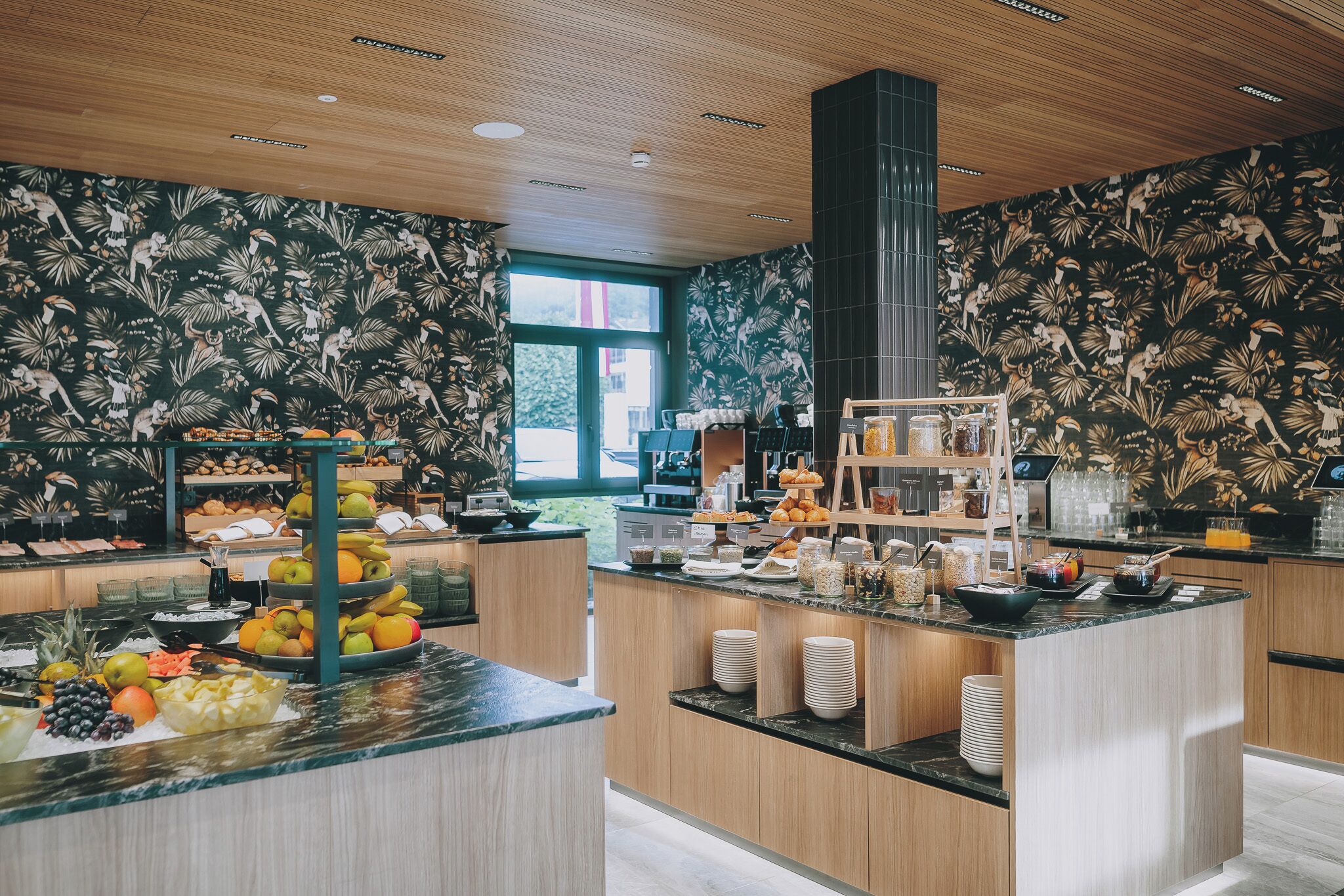 Una cucina moderna con mobili in legno, un'isola centrale con frutta e bevande, e una carta da parati con motivi tropicali.