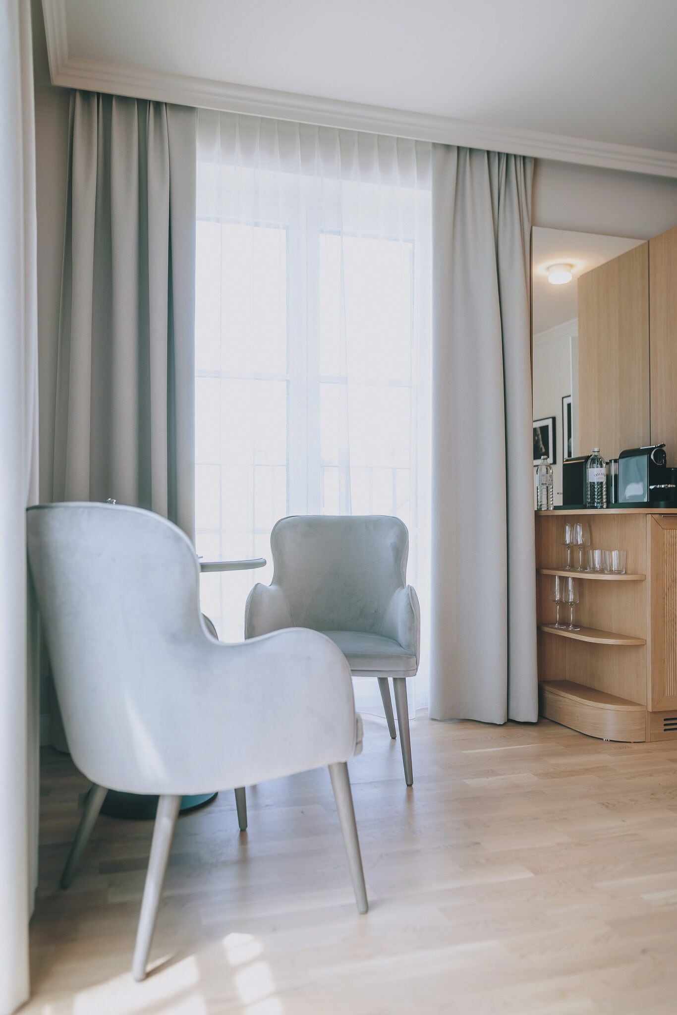 Un saló modern amb un relaxant gris clar, un armari de fusta amb electrodomèstics de cuina i una finestra gran amb cortines translúcides.