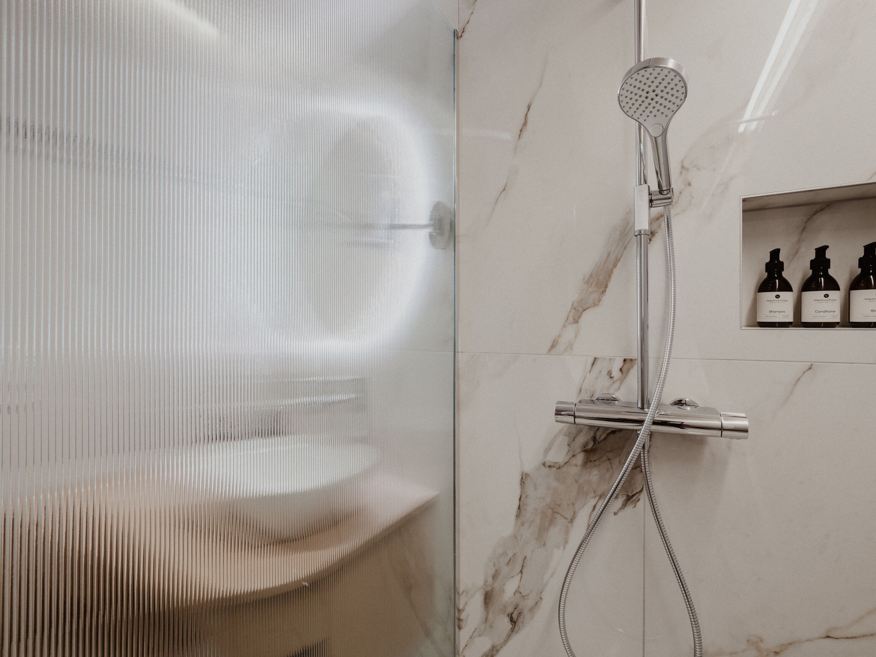 A modern bathroom with a shower, a glass door, and a marble wall. There are three bottles of toiletries on the wall.