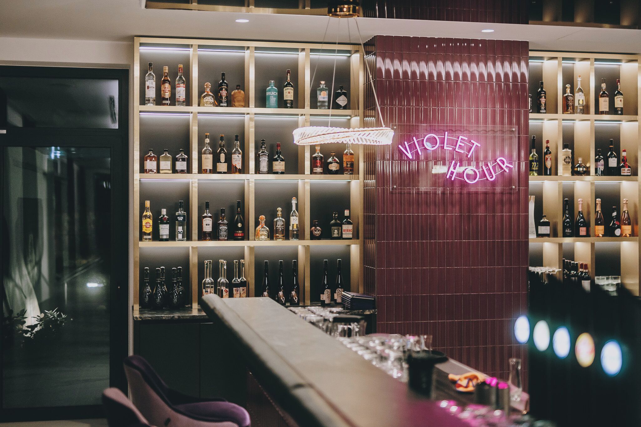 Eine Bar mit Regalen voller Spirituosenflaschen, einem leuchtenden Schild mit der Aufschrift „VIOLET HOUR“ und Barhocker.
