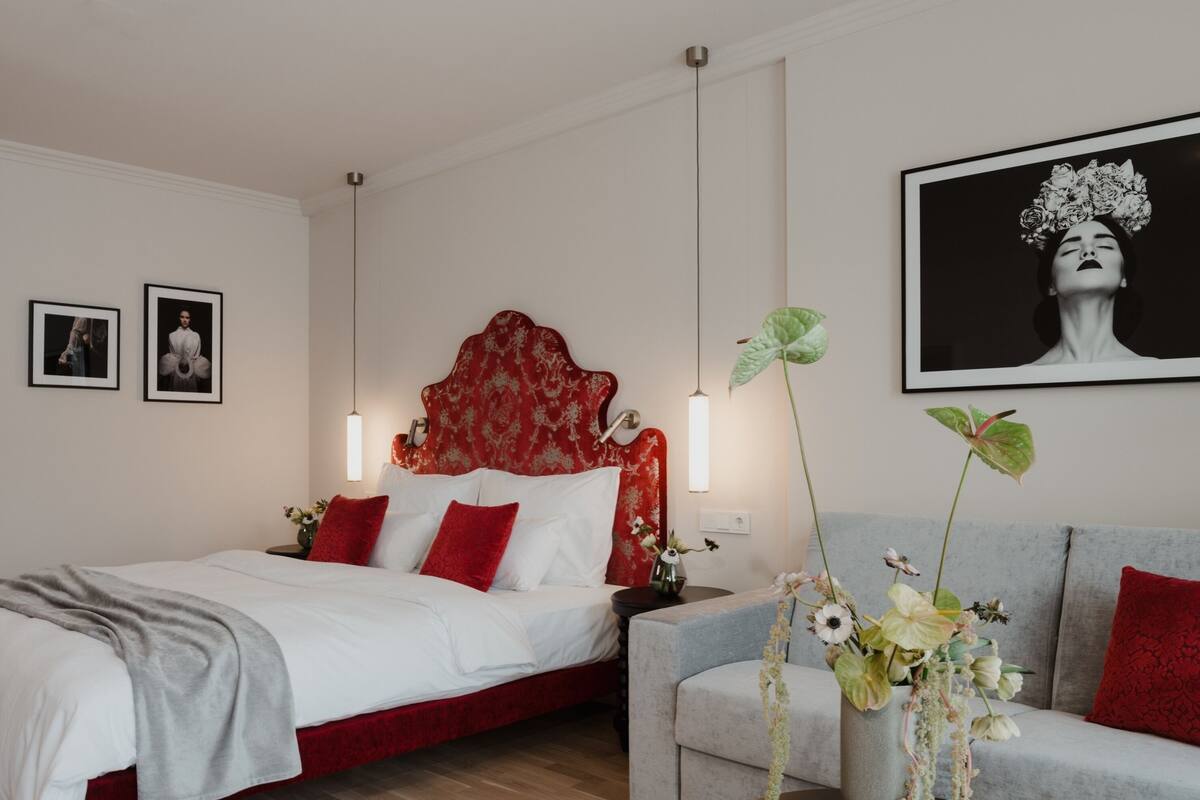 A modern bedroom with a red upholstered bed, a grey sofa, and a large framed artwork on the wall.