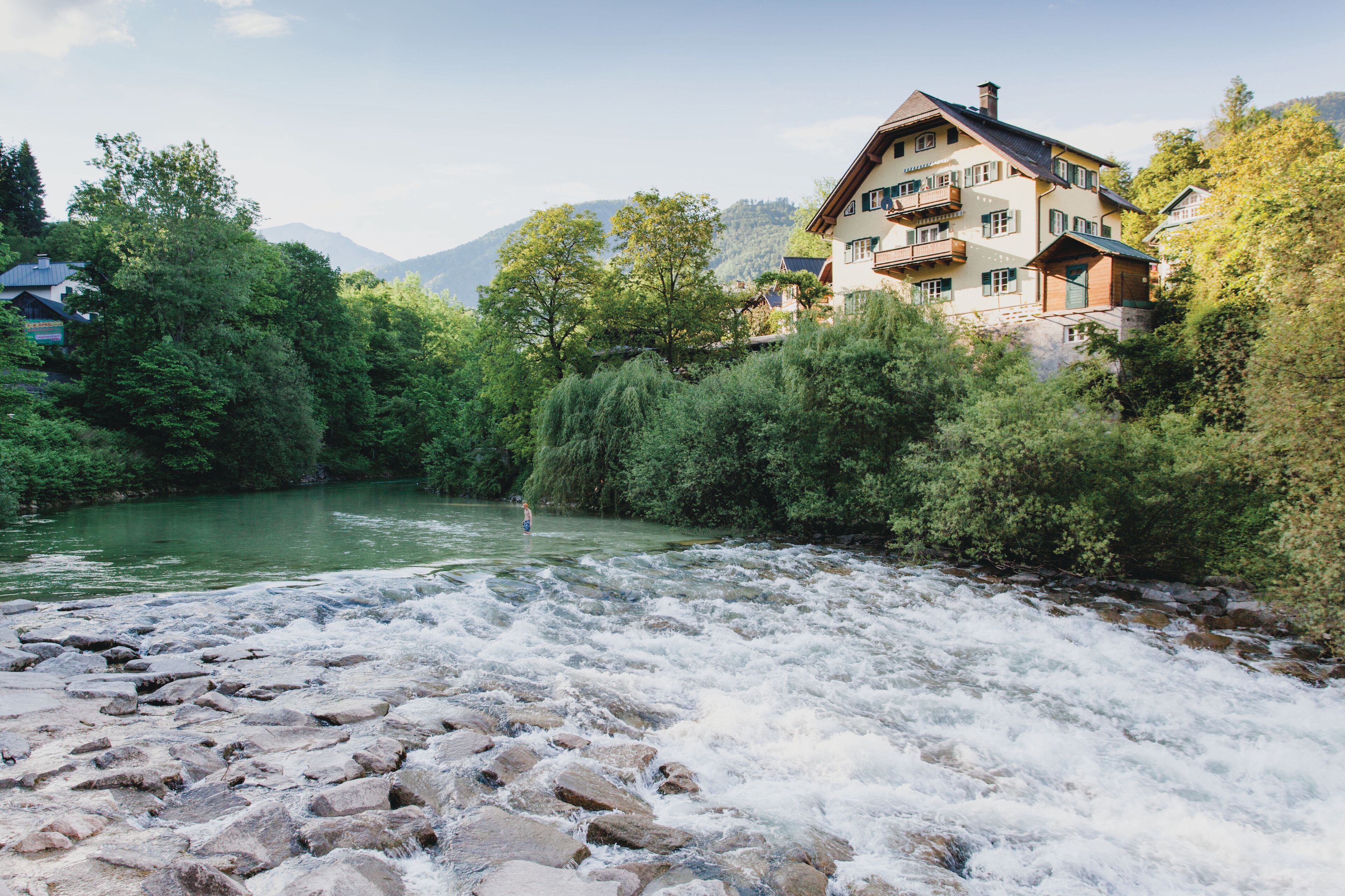 Un hotel junto al río con vista a un río que fluye, rodeado de vegetación exuberante y montañas al fondo.